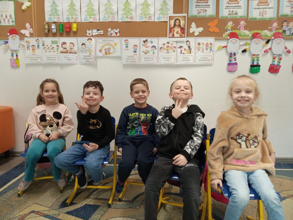 Na zdjęciu pięcioro uśmiechniętych dzieci siedzi na kolorowych krzesłach w przedszkolu. Za nimi widać ścianę ozdobioną edukacyjnymi plakatami i ręcznie robionymi dekoracjami.