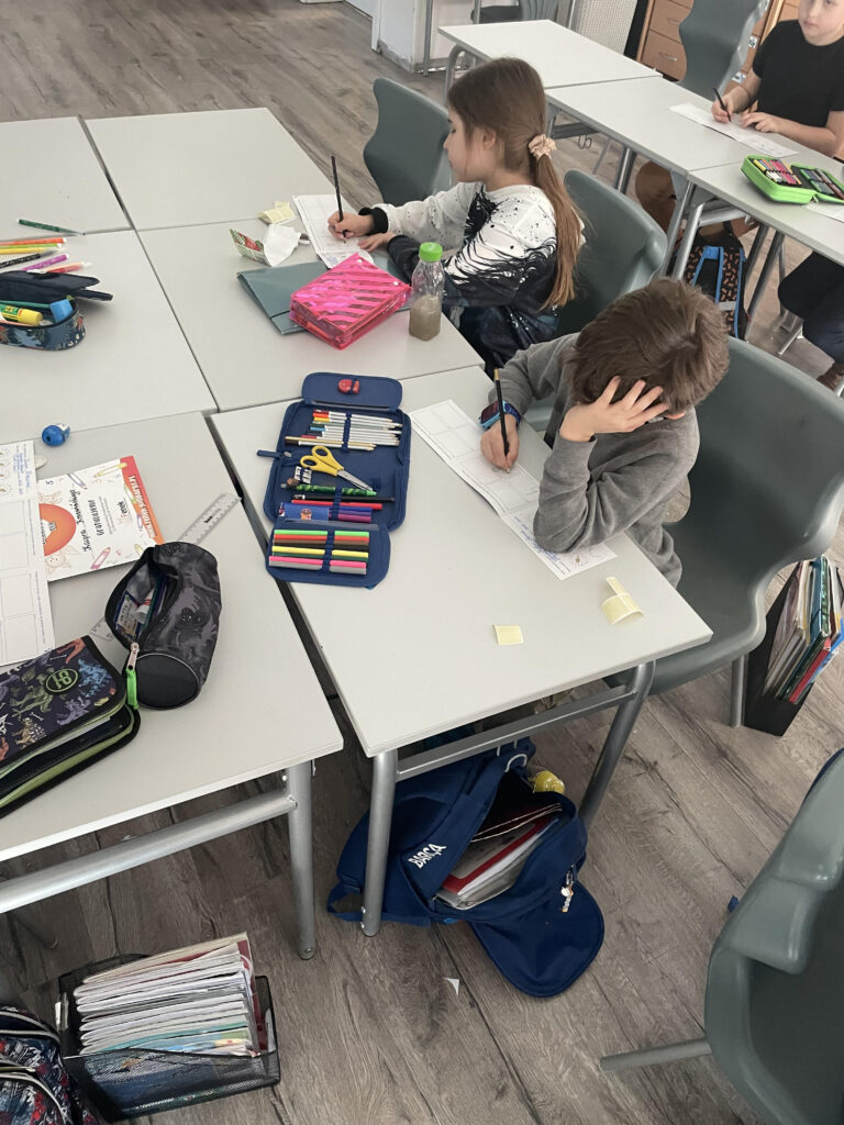 Zdjęcie ukazuje kilkoro dzieci siedzących przy stole w klasie, zajętych rysowaniem przy użyciu kredki i flamastrów z otwartych piórników.