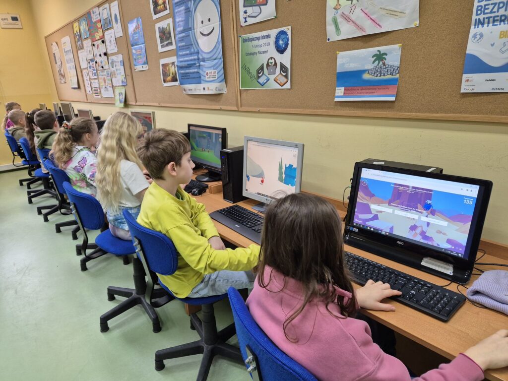 Grupa dzieci siedzi w szkolnej pracowni komputerowej. Skupione są na monitorach, na których wyświetlają się edukacyjne gry lub programy. W tle widać plakaty dotyczące bezpieczeństwa w internecie oraz prace dzieci na ten temat.