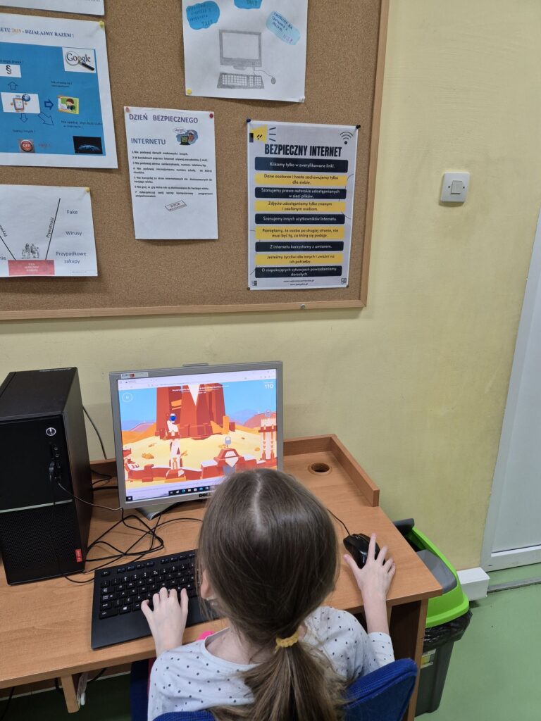 Dziewczynka siedzi przy komputerze w klasie szkolnej, grając w edukacyjną grę komputerową. Na ścianie za nią wisi plakat z zasadami bezpieczeństwa w internecie, który zawiera różne wskazówki i ostrzeżenia.
