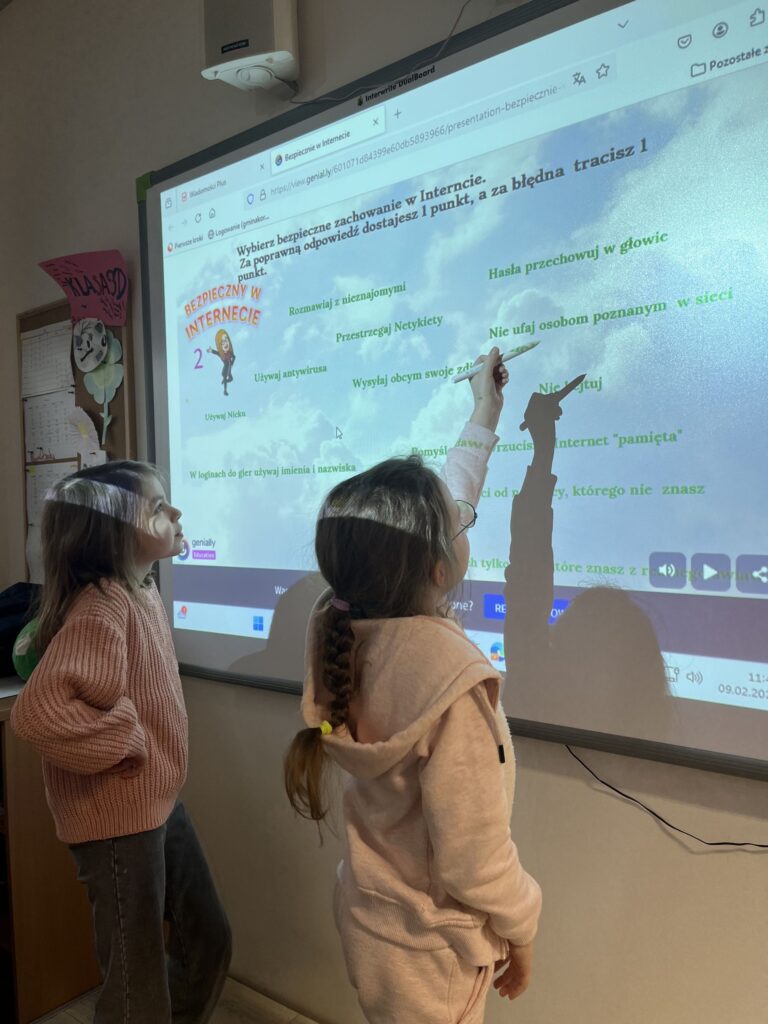 Dwie młode dziewczynki stoją w klasie szkolnej, wskazują na projektor multimedialny, który wyświetla prezentację na temat bezpiecznego zachowania w internecie. Jedna z dziewcząt wskazuje palcem na ekran, gdzie wyświetlane są różne wskazówki dotyczące cyberbezpieczeństwa.