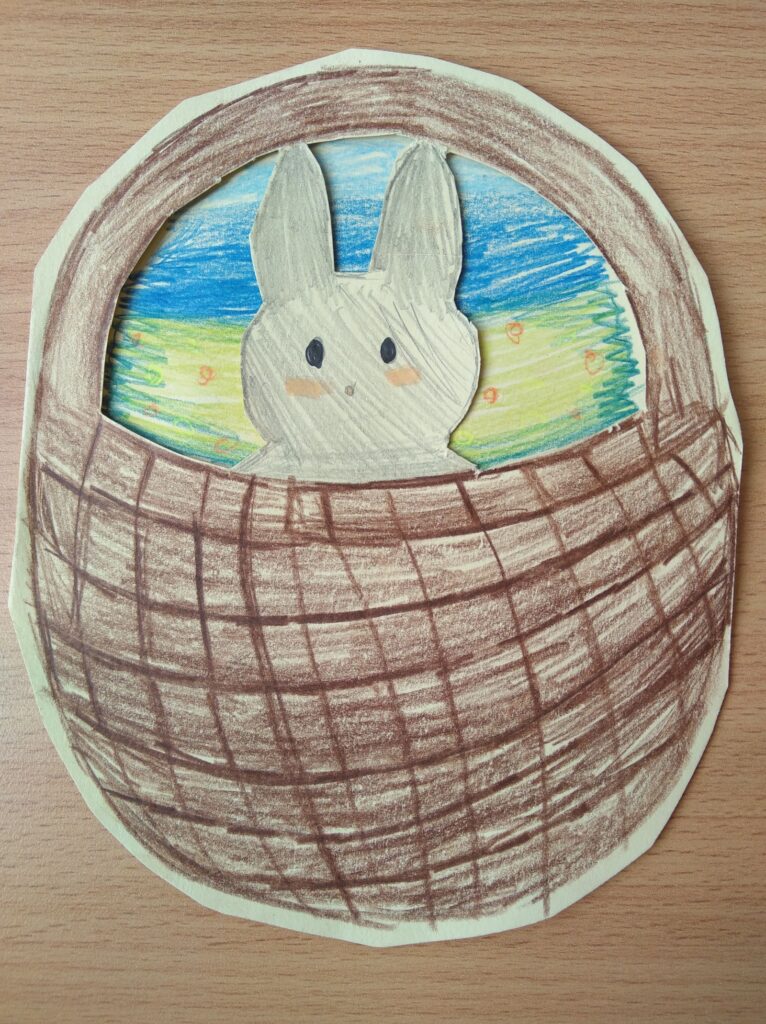 Kartka z rysunkiem przedstawiającym urocze szare króliczki wychylające się z brązowego koszyka. Tło ilustracji jest niebieskie z zielonymi plamami, co sugeruje wiosenną scenerię.