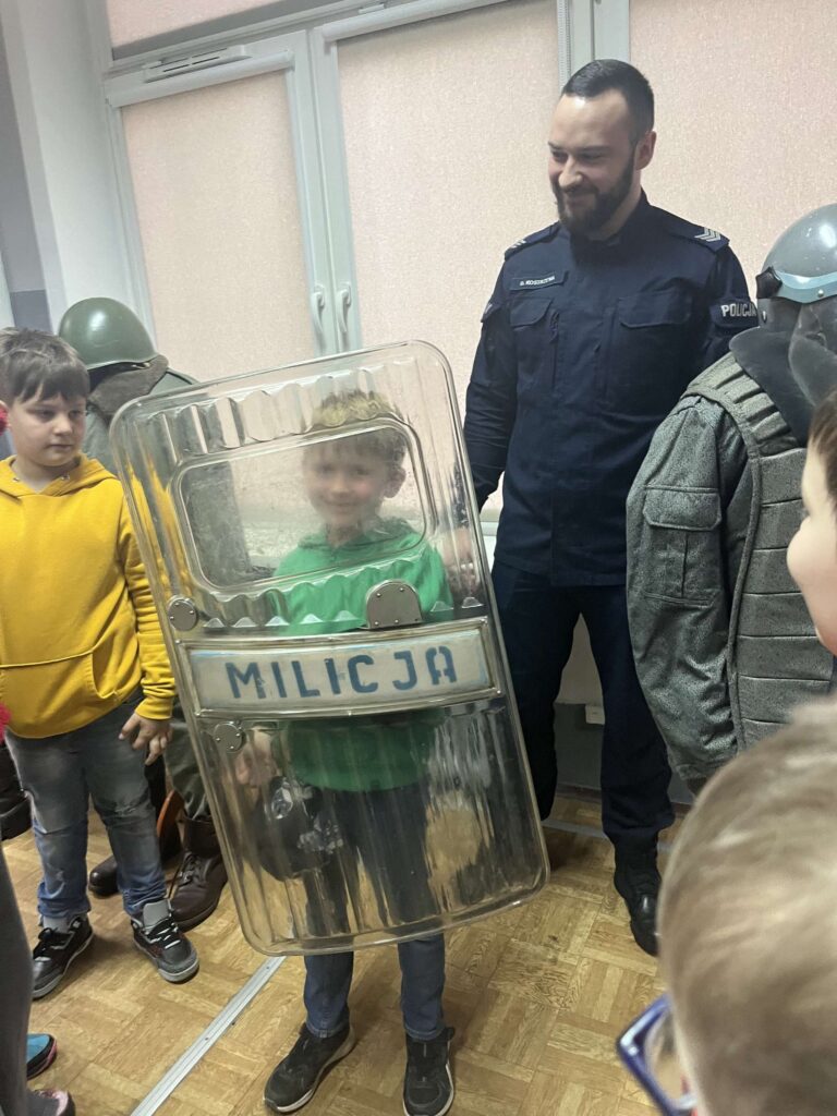 Chłopiec w zielonej bluzie ma na sobie przezroczystą tarczę z napisem "MILICJA" i uśmiecha się, stojąc obok policjanta i wśród innych dzieci.