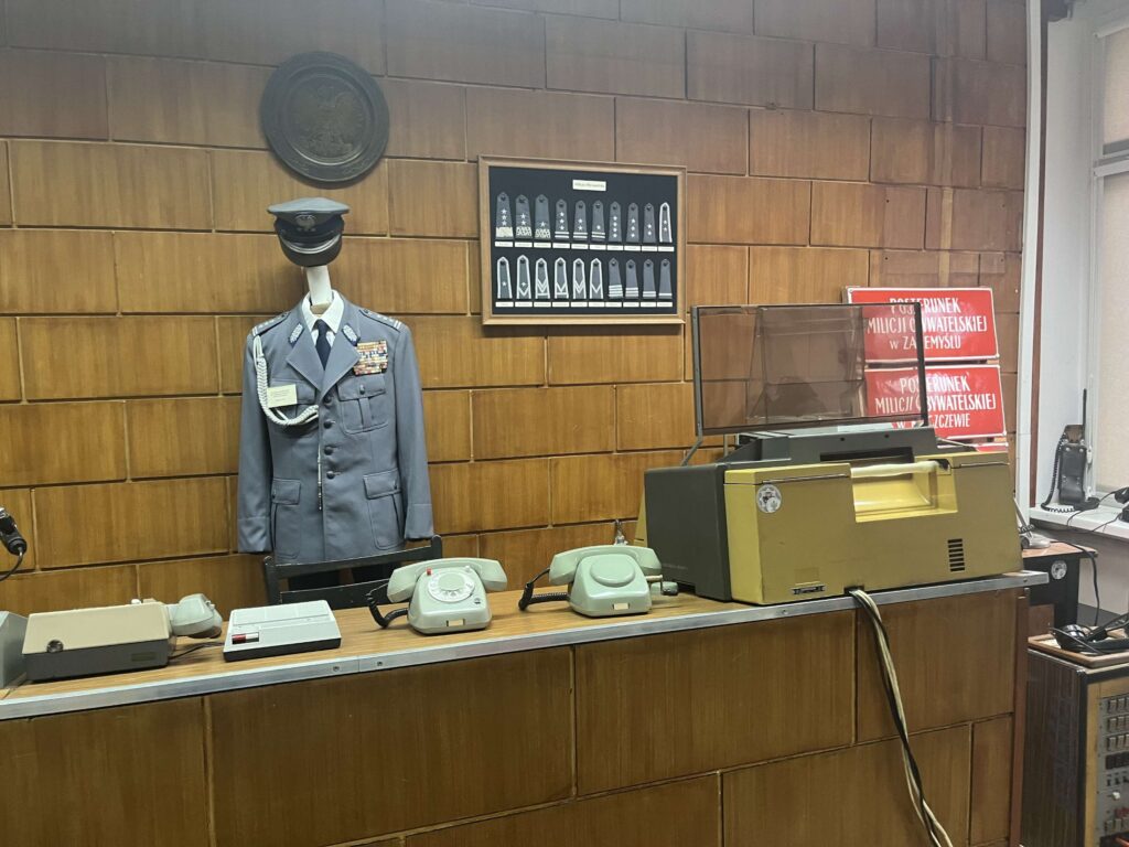 Stary policyjny mundur wystawiony na manekinie, obok starej maszyny do pisania, telefonów i innego sprzętu z czasów minionych.