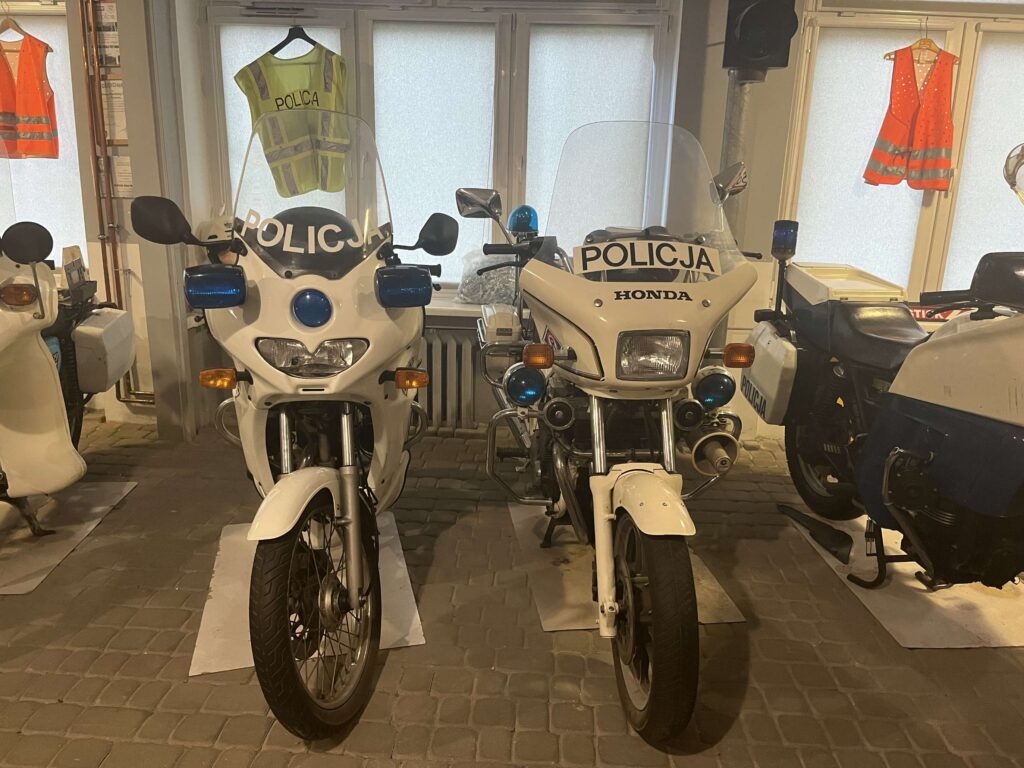 Dwa policyjne motocykle marki Honda stoją wystawione w pomieszczeniu, z policyjnymi kamizelkami odblaskowymi na tle.
