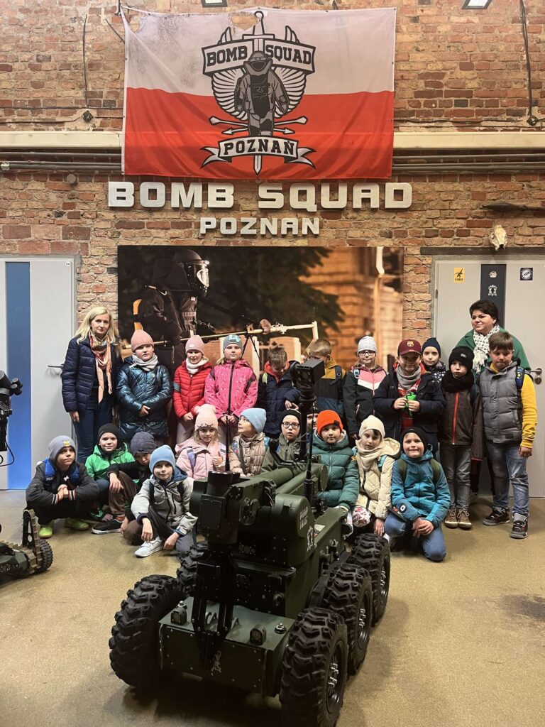 Grupa dzieci stoi przed robotem do rozminowywania bomb, pod wielkim banerem z napisem "BOMB SQUAD POZNAŃ" w pomieszczeniu o ceglanych ścianach.