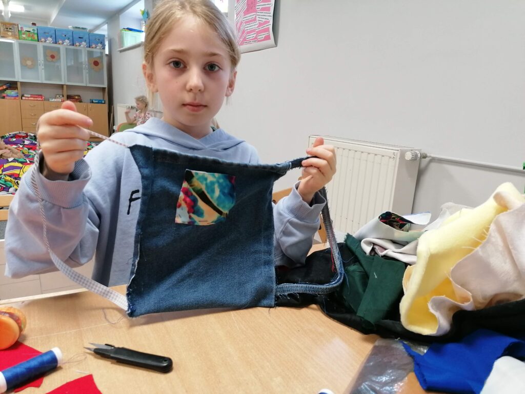  Dziewczynka w niebieskiej bluzie prezentuje ręcznie wykonaną pracę z tkanin, prawdopodobnie torbę, która ma na sobie kolorową łatkę. Inne materiały do szycia są rozłożone na stole wokół niej, a w tle widoczna jest część klasy szkolnej.