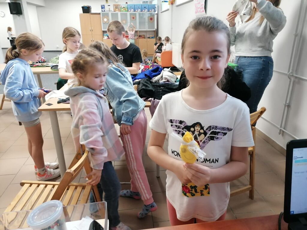  Dziewczynka w białym T-shircie z nadrukiem "Wonder Woman" stoi w klasie, trzymając w rękach żółtą, ręcznie robioną maskotkę. W tle widać inne dzieci i wnętrze klasy.