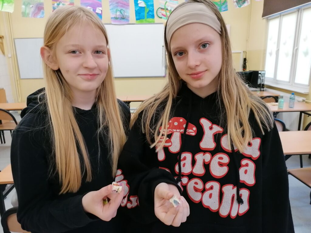 Dwie dziewczynki stoją w klasie szkolnej, prezentując własnoręcznie wykonane przedmioty. Jedna z nich ma na sobie bluzę z napisem "You are a dream", a za nimi widać jasne wnętrze klasy i inne dzieci.
