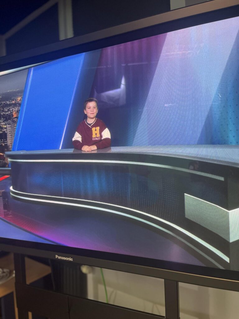 zdjęcie przedstawia chłopca siedzącego przy biurku w studiu telewizyjnym. Na ekranie widoczny jest profesjonalny zestaw do prowadzenia programu informacyjnego.