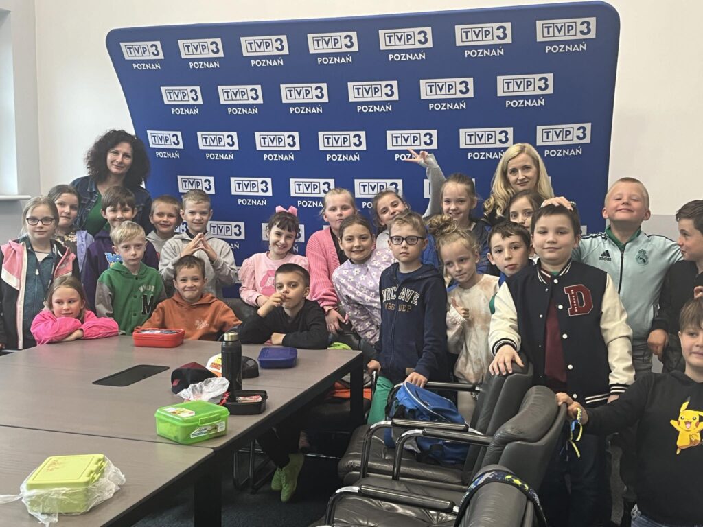  grupa dzieci, nauczycielka oraz baner TVP3 Poznań w tle. Uczniowie i nauczycielka uśmiechają się do aparatu.