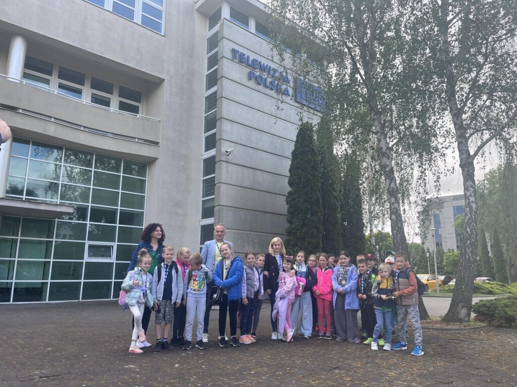 grupowe zdjęcie dzieci i ich opiekunów na tle budynku Telewizji Polskiej w Poznaniu. Wszyscy stoją przed wejściem do budynku.