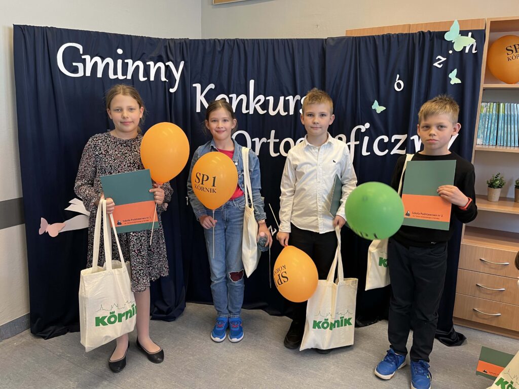 Grupa dzieci trzymających balony stoi w sali przed tablicą z napisem "Gminny konkurs ortograficzny". W dłoniach trzymają torbę z nagrodami i teczkę z dyplomem.